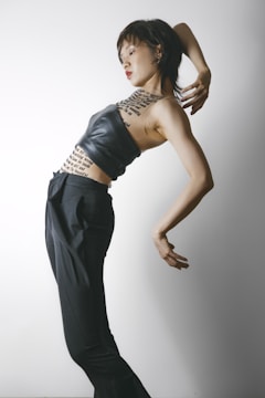 A person is posing artistically against a plain background. They are wearing a black leather top and high-waisted black trousers. Their torso is adorned with text, possibly resembling tattoos or body art. The posture is expressive and dynamic, suggesting movement or dance.