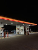 A gas station with brightly lit pumps and a canopy overhead. The scene is mostly empty, with several fueling stations visible. The area is well-lit against the dark night sky, with illuminated signage on the pumps.
