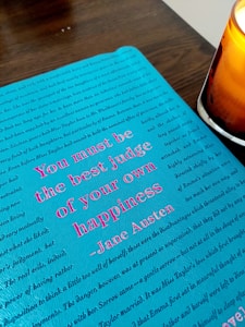 A close-up of a page with a turquoise background featuring a quote in pink text. The quote by Jane Austen reads 'You must be the best judge of your own happiness.' The page is decorated with additional text in a faint dark script. A lit candle with an orange hue is positioned to the right, casting a warm glow.