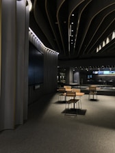 A modern, dimly lit interior space with sleek architectural features. The ceiling design incorporates dark panels with integrated lighting that creates an ambient atmosphere. In the center, there is a set of tables and chairs with wooden finishes on a carpeted floor. In the background, a large screen and additional tables are visible.