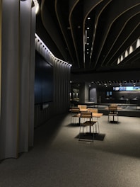 A modern, dimly lit interior space with sleek architectural features. The ceiling design incorporates dark panels with integrated lighting that creates an ambient atmosphere. In the center, there is a set of tables and chairs with wooden finishes on a carpeted floor. In the background, a large screen and additional tables are visible.