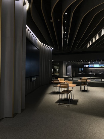 A modern, dimly lit interior space with sleek architectural features. The ceiling design incorporates dark panels with integrated lighting that creates an ambient atmosphere. In the center, there is a set of tables and chairs with wooden finishes on a carpeted floor. In the background, a large screen and additional tables are visible.