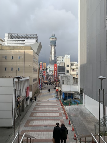 A cityscape with a tall observation tower in the background. Buildings line both sides of the street, which is populated with a few people walking. Bright signs with Japanese characters adorn the street. The sky appears overcast, and the path is lined with orange traffic cones.
