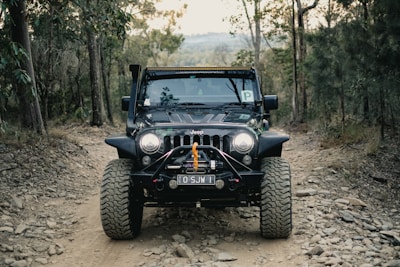 A black Jeep with rugged tires is driving on a rocky dirt path surrounded by trees. The vehicle appears equipped for off-road adventures and features a prominent front bumper with a winch. The landscape suggests a forest environment, with various trees and bushes lining the path.