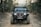 A black Jeep with rugged tires is driving on a rocky dirt path surrounded by trees. The vehicle appears equipped for off-road adventures and features a prominent front bumper with a winch. The landscape suggests a forest environment, with various trees and bushes lining the path.
