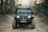 A black Jeep with rugged tires is driving on a rocky dirt path surrounded by trees. The vehicle appears equipped for off-road adventures and features a prominent front bumper with a winch. The landscape suggests a forest environment, with various trees and bushes lining the path.