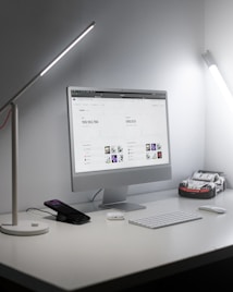 A modern, minimalistic workspace featuring a sleek desktop computer displaying statistical graphs on a screen, accompanied by a keyboard and mouse on a white desk. Two desk lamps provide ample lighting. A smartphone is placed on a stand, and a toy car model is situated next to the computer.