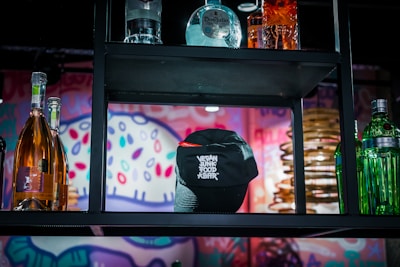 A bar shelf displaying various liquor bottles including tequila and whiskey. A black cap with text on it is prominently placed at the center. The background features a colorful, abstract mural with bright patterns and shapes.