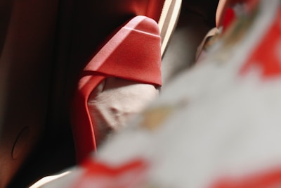 A close-up image showcasing the elegant curve of a high-heeled red shoe. Soft shadows and light create a dramatic and stylish atmosphere, with parts of the shoe blurred in the foreground, suggesting a sense of movement or depth.