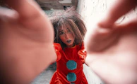 A person dressed as a clown is reaching towards the camera with a distressed expression. The clown is wearing a bright red costume with large blue pompoms, and their hair is messy, adding to the dramatic effect. The background is a textured wall, giving an impression of an urban or abandoned setting.