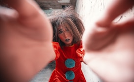 A person dressed as a clown is reaching towards the camera with a distressed expression. The clown is wearing a bright red costume with large blue pompoms, and their hair is messy, adding to the dramatic effect. The background is a textured wall, giving an impression of an urban or abandoned setting.
