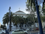 A building with a large dome and cross at the top, identified as the First Baptist Church. It is surrounded by tall palm trees and other greenery. Several cars are parked along the street, and there are streetlights and a no parking sign visible.