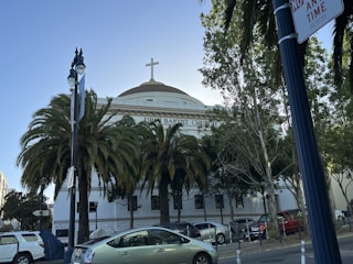 A building with a large dome and cross at the top, identified as the First Baptist Church. It is surrounded by tall palm trees and other greenery. Several cars are parked along the street, and there are streetlights and a no parking sign visible.