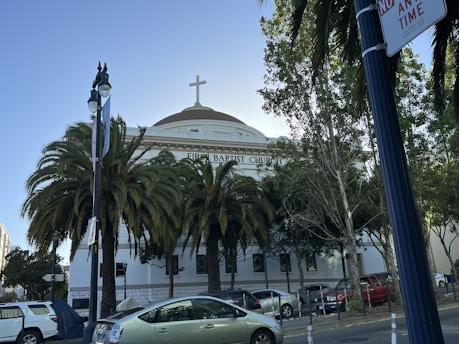 A building with a large dome and cross at the top, identified as the First Baptist Church. It is surrounded by tall palm trees and other greenery. Several cars are parked along the street, and there are streetlights and a no parking sign visible.