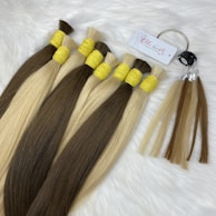 Bundles of straight hair extensions in two colors, dark brown and light blonde, secured with yellow bands. A metal ring holds sample strands with a tag attached, all placed on a soft, white furry surface.