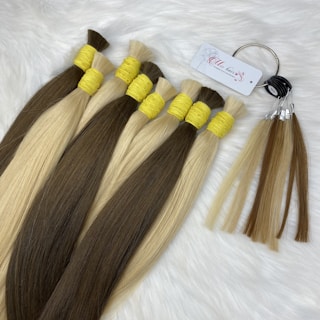 Bundles of straight hair extensions in two colors, dark brown and light blonde, secured with yellow bands. A metal ring holds sample strands with a tag attached, all placed on a soft, white furry surface.