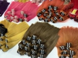 Multiple bundles of hair in a variety of vibrant colors are neatly arranged on a white surface. The colors include pink, red, purple, yellow, and brown. Each bundle is tied securely and appears to be straight and smooth, with the strands lying parallel to one another.