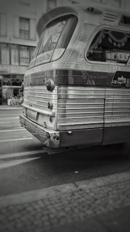A vintage bus with textured metal surface and rounded edges is positioned on a city street. The bus has a retro design featuring rivet details and a rounded rear window. The surroundings suggest an urban environment with buildings and road markings visible.