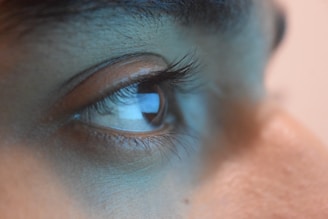 Close-up of a human eye with visible eyelashes and eyebrow, showcasing the texture of the skin around the eye. The reflection of light is visible in the cornea, highlighting the eye's detail.