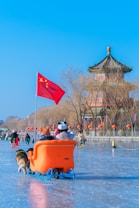 A winter scene with people enjoying ice skating. An orange sled with two passengers, one wearing a panda hat, is being pushed by a dog. A large Chinese flag waves behind the sled. Traditional Chinese architecture, characterized by a pagoda-style building, appears in the background. Red lanterns decorate the area, and bare trees line the scene. The ice reflects the bright blue sky.
