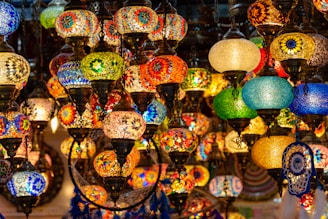 An assortment of colorful, intricately designed mosaic lamps hang closely together. The lamps feature patterns made from vibrant glass pieces in various shapes and sizes, creating a warm and inviting ambiance. The fusion of colors includes blues, reds, greens, and yellows, with some lamps featuring traditional motifs.