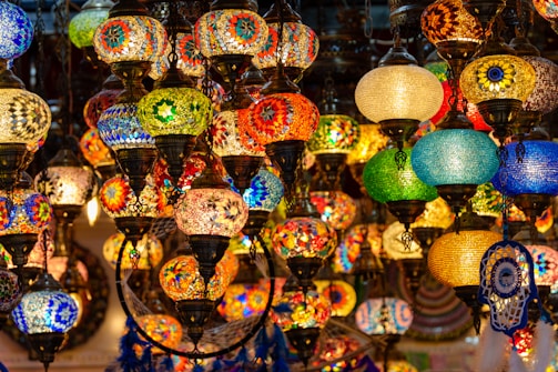 An assortment of colorful, intricately designed mosaic lamps hang closely together. The lamps feature patterns made from vibrant glass pieces in various shapes and sizes, creating a warm and inviting ambiance. The fusion of colors includes blues, reds, greens, and yellows, with some lamps featuring traditional motifs.