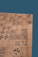 A wooden surface features a series of square panels with intricate laser-cut, geometric and floral patterns. The background is a solid teal color.