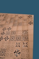 A wooden surface features a series of square panels with intricate laser-cut, geometric and floral patterns. The background is a solid teal color.