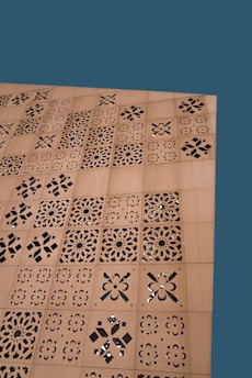 A wooden surface features a series of square panels with intricate laser-cut, geometric and floral patterns. The background is a solid teal color.