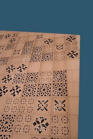 A wooden surface features a series of square panels with intricate laser-cut, geometric and floral patterns. The background is a solid teal color.