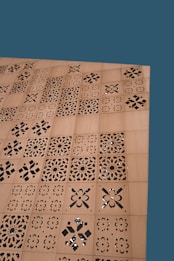 A wooden surface features a series of square panels with intricate laser-cut, geometric and floral patterns. The background is a solid teal color.