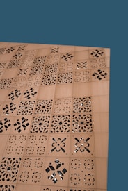 A wooden surface features a series of square panels with intricate laser-cut, geometric and floral patterns. The background is a solid teal color.