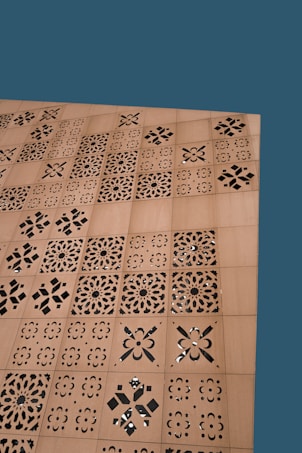 A wooden surface features a series of square panels with intricate laser-cut, geometric and floral patterns. The background is a solid teal color.