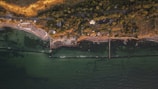 An aerial view of a coastal area with a green sea, a shoreline with sandy beaches, and dense vegetation. A road runs parallel to the coast, with several buildings and structures among the trees. Breakwaters extend into the sea, creating sheltered areas along the coast.