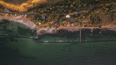 An aerial view of a coastal area with a green sea, a shoreline with sandy beaches, and dense vegetation. A road runs parallel to the coast, with several buildings and structures among the trees. Breakwaters extend into the sea, creating sheltered areas along the coast.