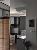 A modern kitchen features sleek cabinetry with light wood and dark finishes, a black range hood, and a gas stove. A counter area includes small kitchen appliances and utensils, with a unique clock on the wall. A decorative wall sign adds a personal touch, and a vase with flowers sits nearby, enhancing the decor.