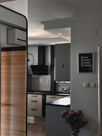 A modern kitchen features sleek cabinetry with light wood and dark finishes, a black range hood, and a gas stove. A counter area includes small kitchen appliances and utensils, with a unique clock on the wall. A decorative wall sign adds a personal touch, and a vase with flowers sits nearby, enhancing the decor.