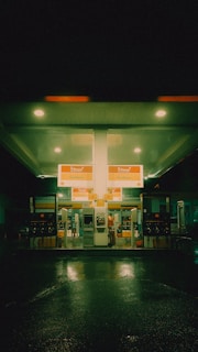 A gas station at night with a neon green glow fills the scene. The station is illuminated by overhead lights, reflecting on the wet pavement. Bright signage indicates fuel options, and there is a person at one of the pumps.