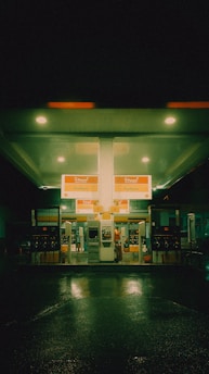 A gas station at night with a neon green glow fills the scene. The station is illuminated by overhead lights, reflecting on the wet pavement. Bright signage indicates fuel options, and there is a person at one of the pumps.