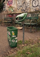 A green trash can with various stickers is prominently displayed, with no lid, placed next to a green metal support. Two green benches with graffiti are in the background, along with a wall covered in colorful graffiti. The path is surrounded by patchy grass and dry leaves.