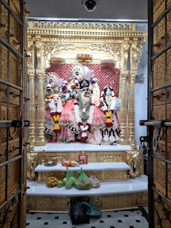 An ornate altar featuring intricately dressed deities adorned with vibrant garlands and positioned against a golden and red backdrop. The altar is framed by an elaborately carved golden structure, and offerings of fruits and other items are placed on a white shelf below.
