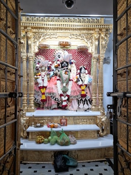 An ornate altar featuring intricately dressed deities adorned with vibrant garlands and positioned against a golden and red backdrop. The altar is framed by an elaborately carved golden structure, and offerings of fruits and other items are placed on a white shelf below.