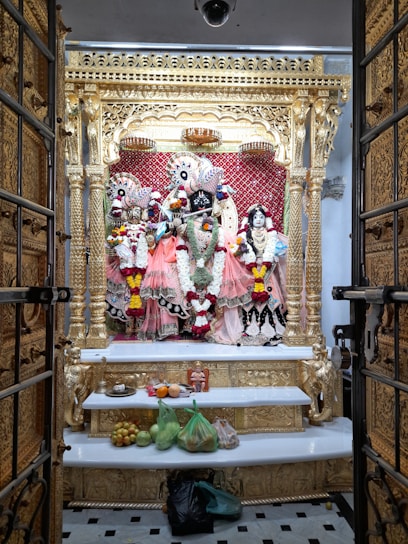 An ornate altar featuring intricately dressed deities adorned with vibrant garlands and positioned against a golden and red backdrop. The altar is framed by an elaborately carved golden structure, and offerings of fruits and other items are placed on a white shelf below.
