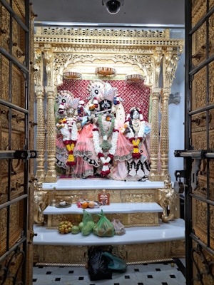 An ornate altar featuring intricately dressed deities adorned with vibrant garlands and positioned against a golden and red backdrop. The altar is framed by an elaborately carved golden structure, and offerings of fruits and other items are placed on a white shelf below.