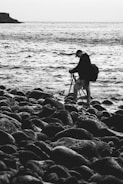 A photographer stands on a rocky shoreline, setting up a tripod while facing the ocean. The scene is captured in black and white, emphasizing the texture of the rocks and the movement of the water.