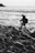 A photographer stands on a rocky shoreline, setting up a tripod while facing the ocean. The scene is captured in black and white, emphasizing the texture of the rocks and the movement of the water.
