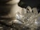 A transparent crystal lotus flower sculpture rests on a textured stone surface next to a marble vase. Light reflects off the crystal, creating subtle rainbow patterns and highlighting the intricate facets of the sculpture.