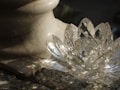A transparent crystal lotus flower sculpture rests on a textured stone surface next to a marble vase. Light reflects off the crystal, creating subtle rainbow patterns and highlighting the intricate facets of the sculpture.
