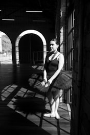 A ballerina stands in a bright, sunlit area with dramatic shadows cast across a wooden floor. She wears a black leotard with a tutu and her hair is neatly styled. The background features large arched windows and brick walls, adding to the industrial setting.