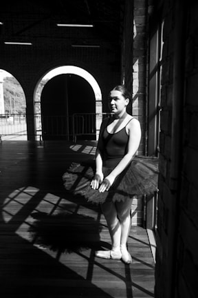 A ballerina stands in a bright, sunlit area with dramatic shadows cast across a wooden floor. She wears a black leotard with a tutu and her hair is neatly styled. The background features large arched windows and brick walls, adding to the industrial setting.